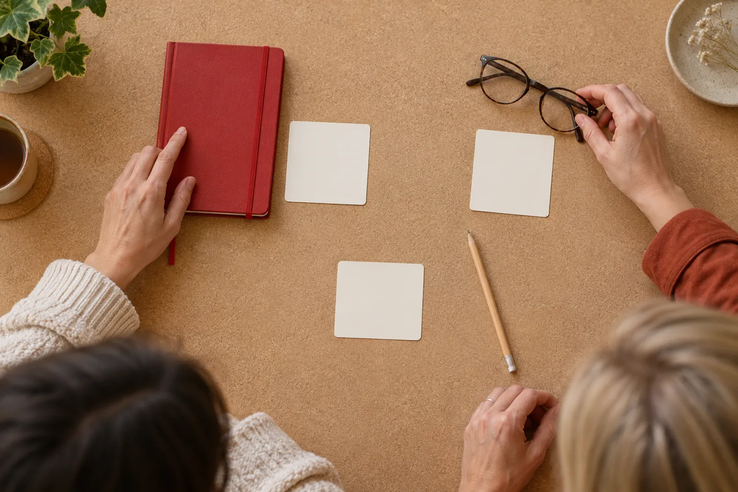 Two hands arranging a red notebook and glasses beside blank cards on a cork surface