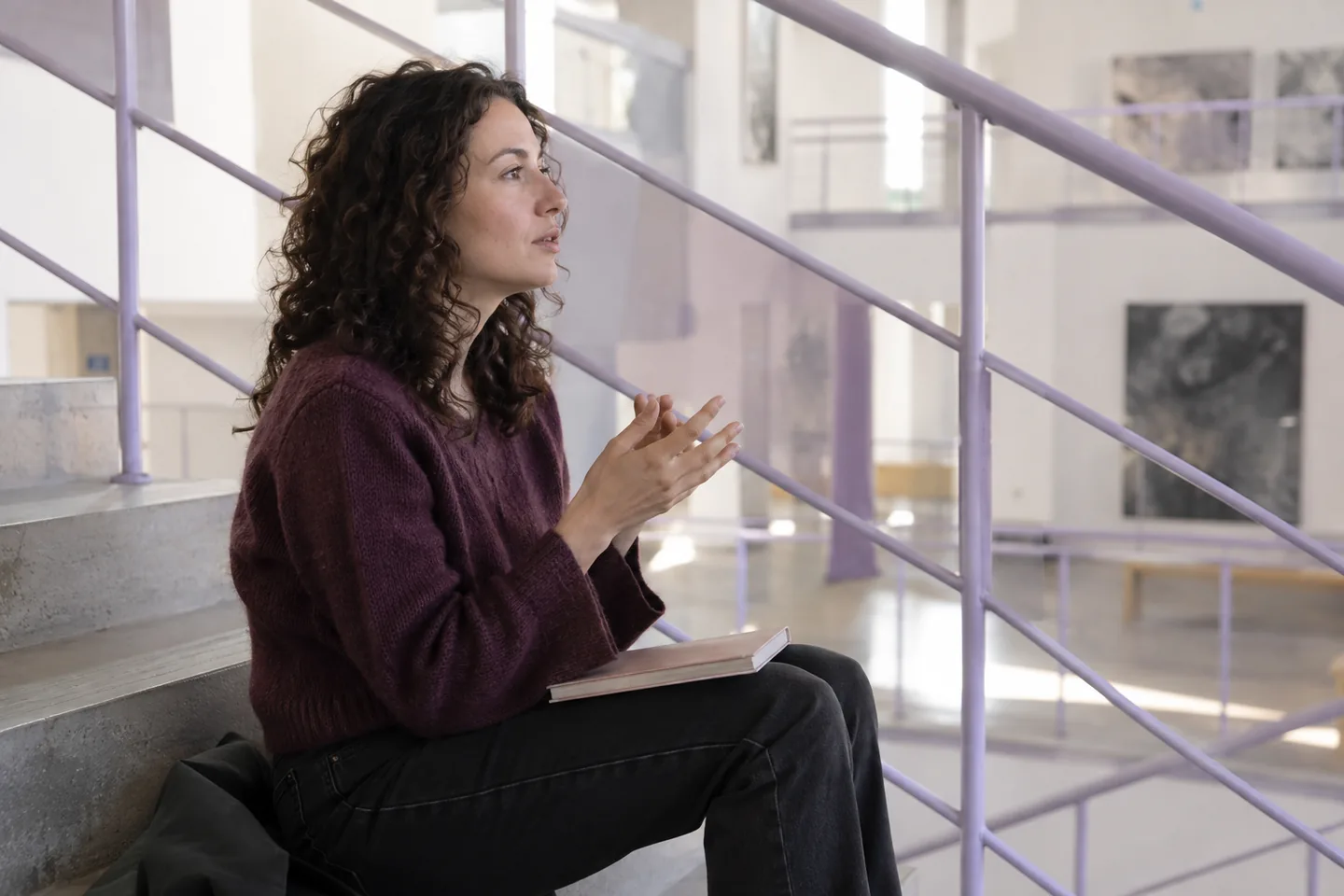 A European woman rehearses a sentence with a closed booklet on an arts-center staircase landing