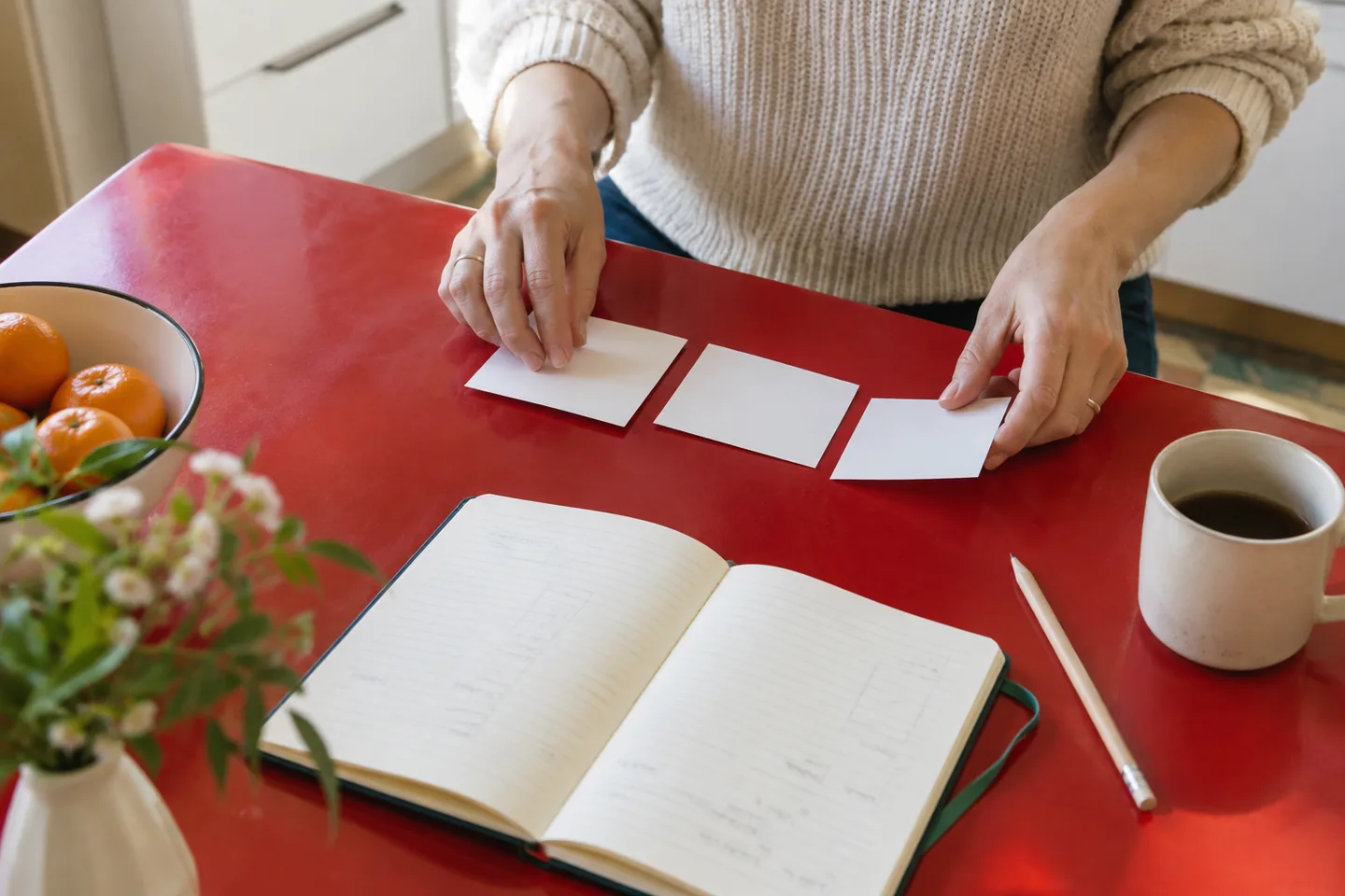 Hands sort three blank task cards beside a notebook on a red kitchen table