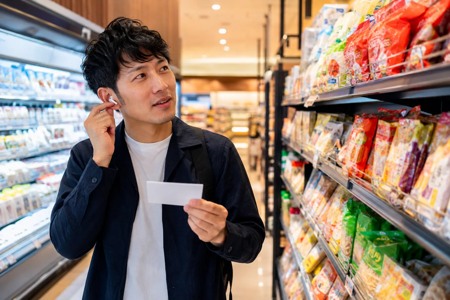 Adult learner listening to katakana loanwords while comparing everyday products in a bright Japanese supermarket aisle